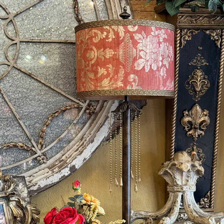 A vintage-style table lamp with a coral pink damask patterned drum shade and gold trim, set on a dark wooden mantel next to a decorative floral arrangement and an ornate white sculpture.