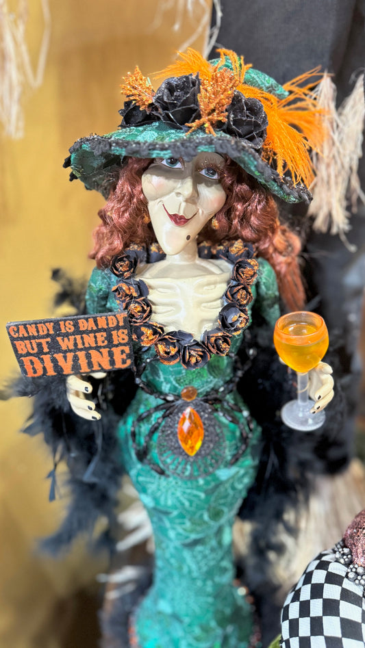 A decorative figure of a green mermaid-like character with a black top hat and a sign that reads 'Candy Wine is Divine'. The character is holding a glass of wine.