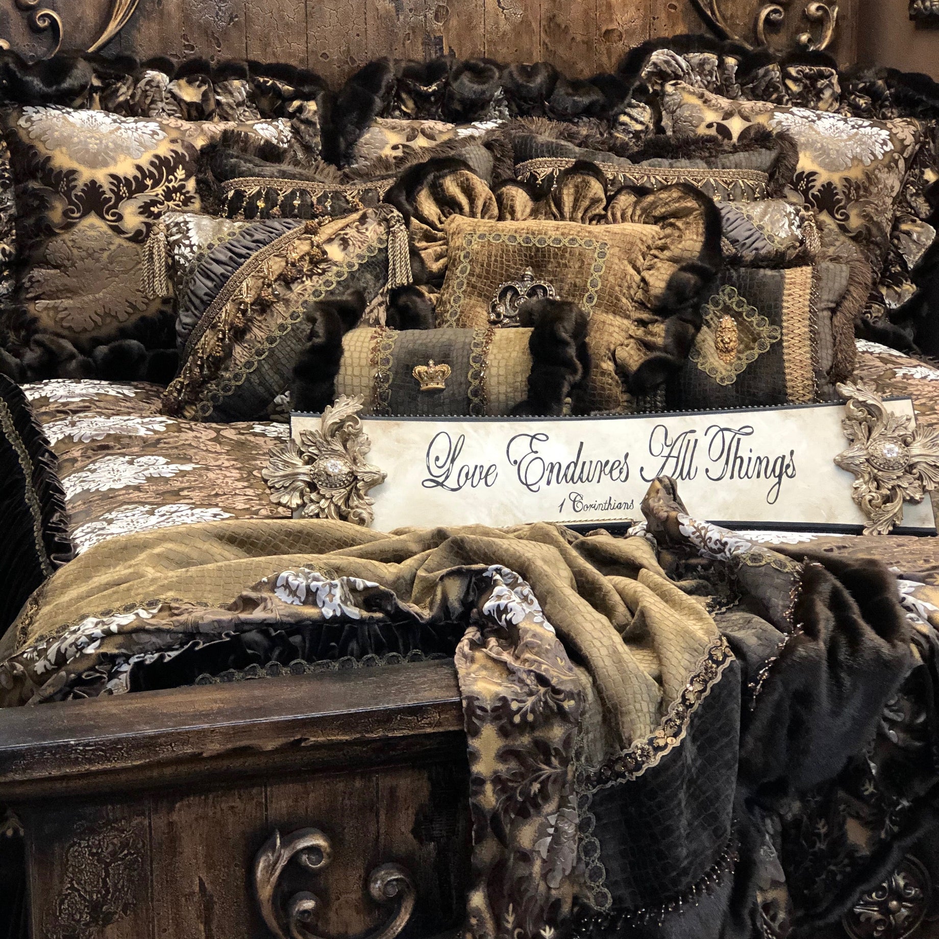 A close-up of an ornate vintage-style bed with heavy drapery, ruffled pillows, and a sign reading 'Love Endures All Things' on a dark wood headboard and footboard.