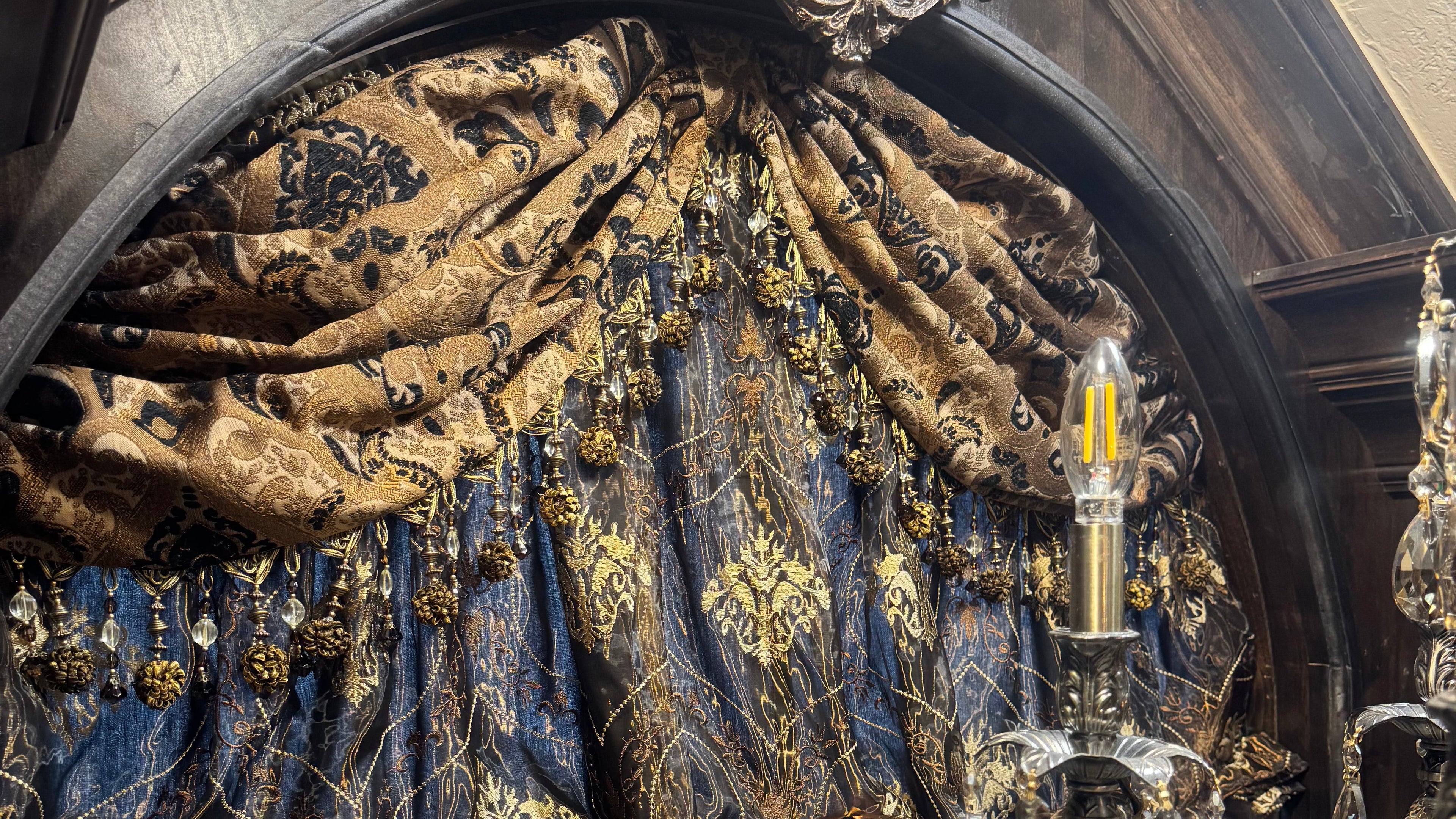 An ornate theatrical curtain with heavy draped fabric in deep blue and gold, featuring intricate embroidery, gold fringe, and a decorative gold crest above an arched opening.