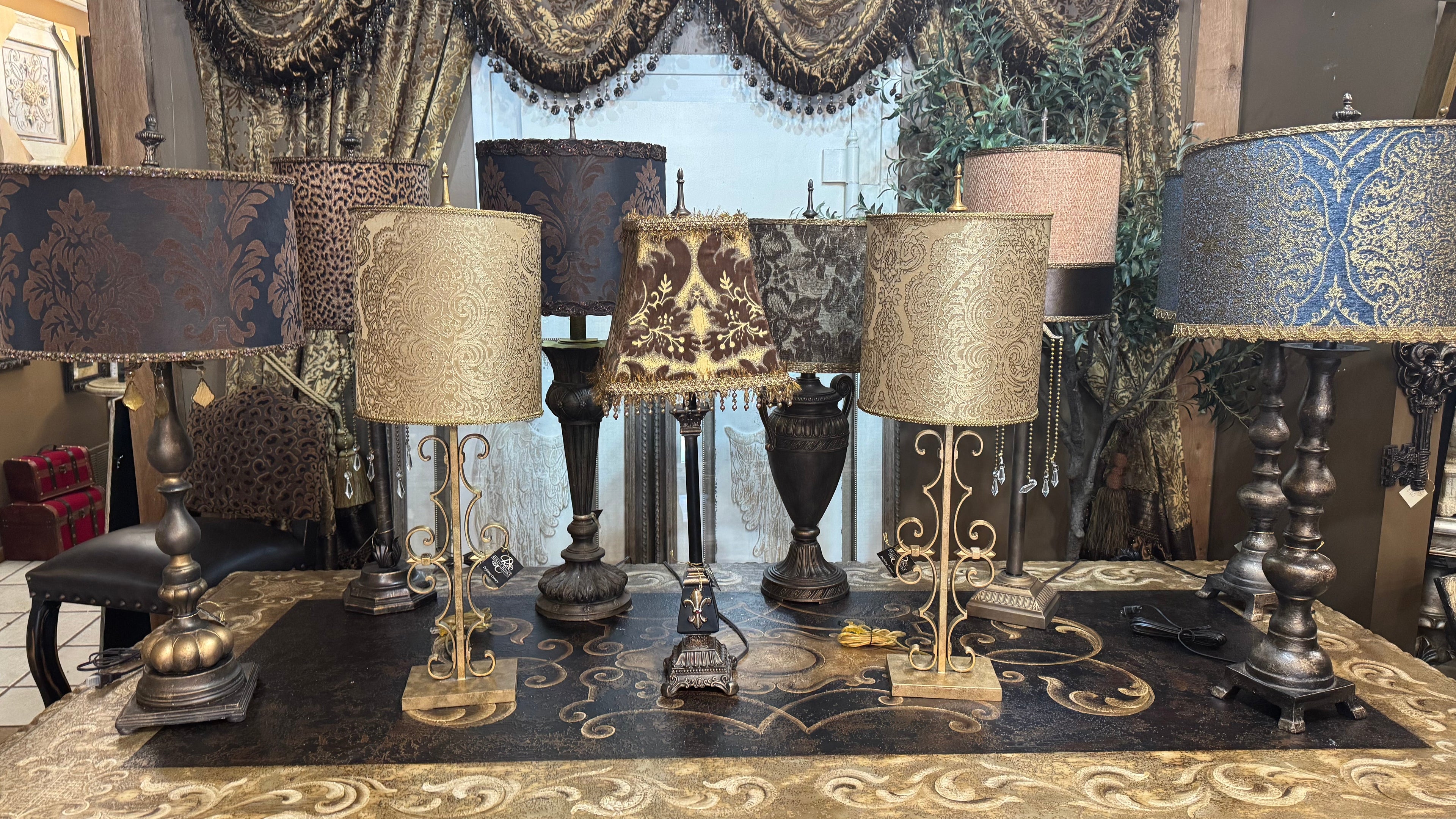 A collection of ornate table lamps displayed on a carved console table in a showroom. The lamps feature various patterned fabric shades in blue, gold, and beige, paired with dark bronze and gold metal bases.