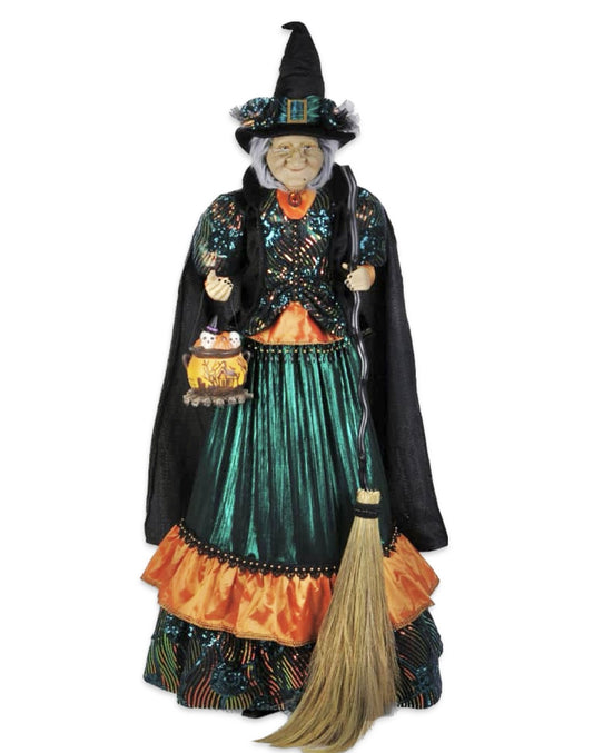 Life size lighted Theodora Witch figurine with a green dress, orange and yellow apron, black hat, and holding a broom. She has a happy facial expression and is dressed for Halloween.