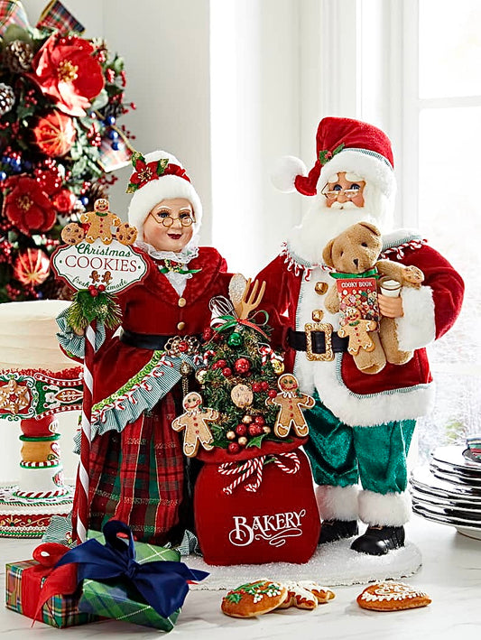 A Lighted Claus Cookie Bakery display featuring figurines of Santa and a Mrs. Claus, with the former holding a teddy bear and a sign, and the latter wearing a red and green plaid dress.